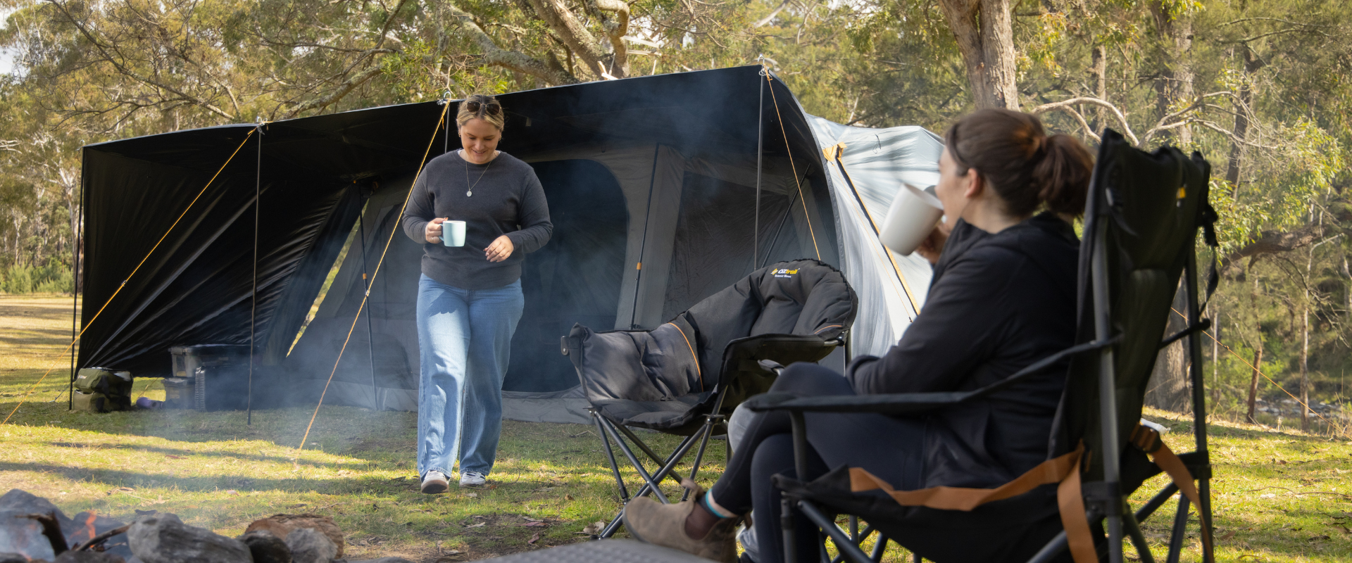 Two campers relaxing with hot drinks beside a large tent, with morning smoke drifting from the campfire.