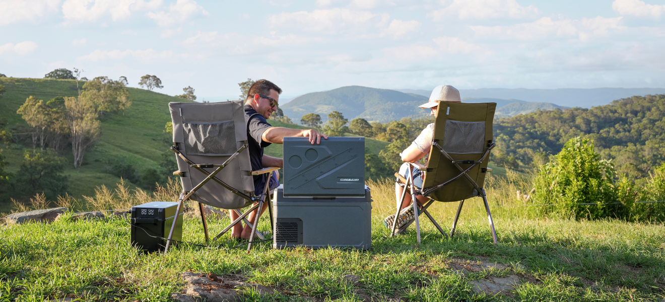 A man and woman relax on a grassy hill in camping chairs, a 30L Companion fridge and power station beside them, enjoying the view