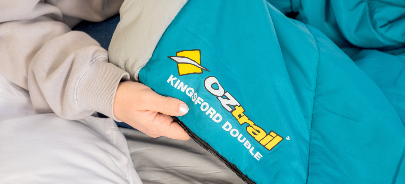 A person wearing a beige hoodie is holding the zipper of a teal Oztrail Kingsford Double sleeping bag.