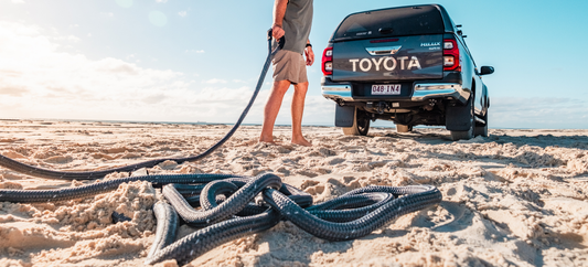 Person standing by a Toyota Hilux on a sandy beach holding a kinetic rope laid out in front of the vehicle.