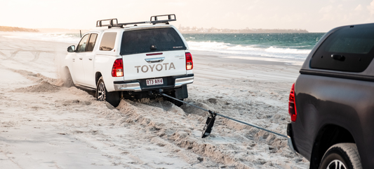 White Toyota Hilux stuck in beach sand being recovered by another vehicle with ocean in background.