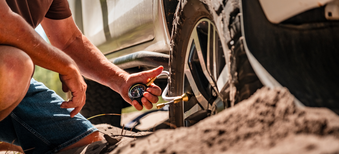 A person is checking the tire pressure of an off-road vehicle with a gauge. The vehicle is on sandy terrain.