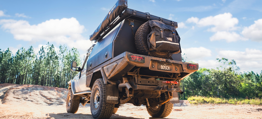 Modified pickup truck with canopy and rear spare tyre parked on dirt track near green forest.