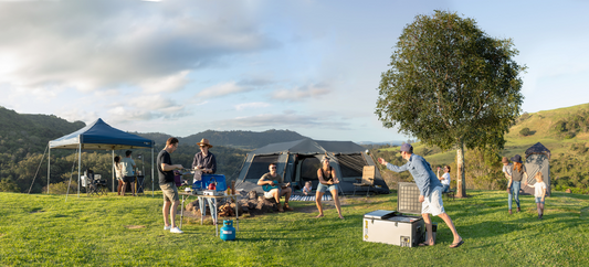Group camping on a sunny hillside, cooking, playing games and relaxing beside tents and a large tree.