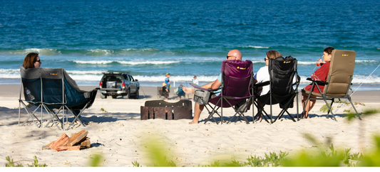 Campers relaxing in folding chairs around a beach fire pit, facing the ocean with waves and a parked 4WD.