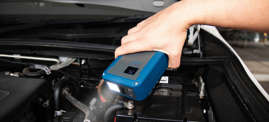 Hand holding a blue Rover 3600A jump starter over a car battery, with digital display and flashlight on.