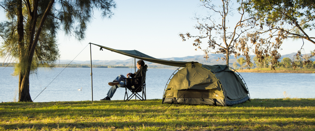 Camper relaxing under a tarp beside a green swag by a lakeside, with trees and distant hills in the background at sunset.