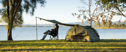 Camper relaxing under a tarp beside a green swag by a lakeside, with trees and distant hills in the background at sunset.