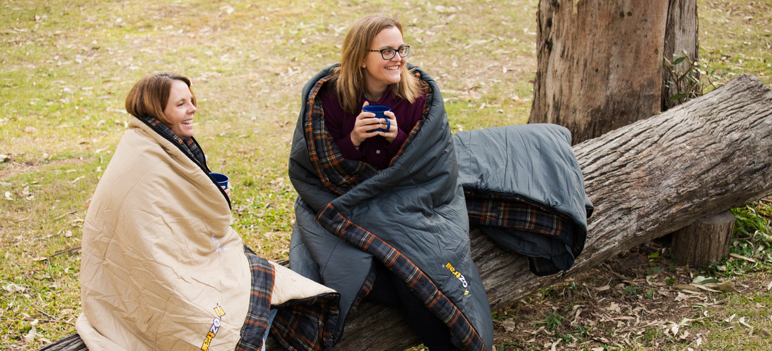 Two people wrapped in sleeping bags sitting on a fallen log, holding mugs at a campsite.