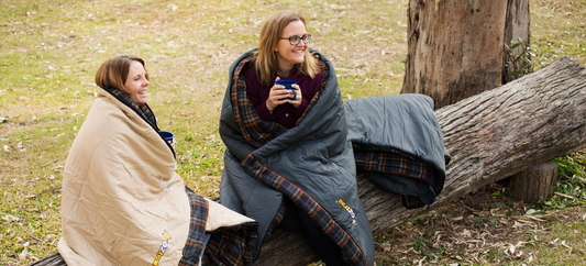 Two people wrapped in sleeping bags sitting on a fallen log, holding mugs at a campsite.
