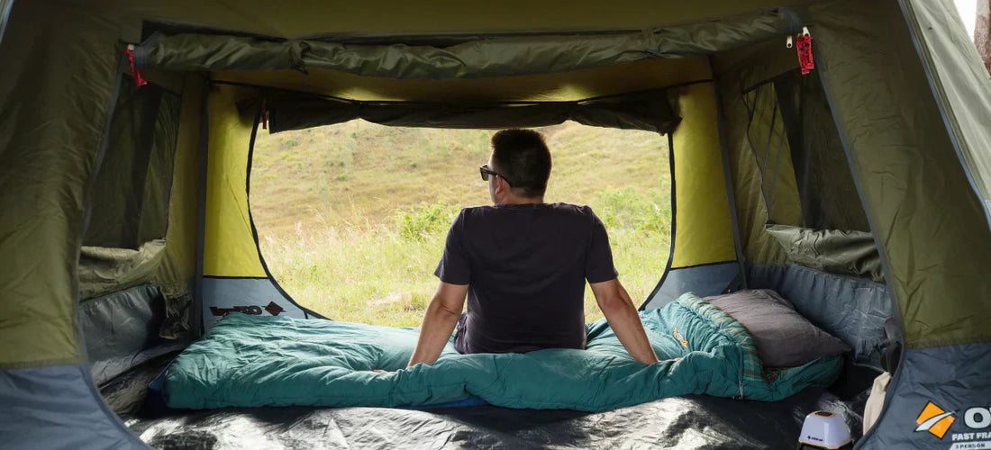 Person relaxing inside an OZtrail Fast Frame tent, sitting on a sleeping bag and looking out toward a grassy hillside.