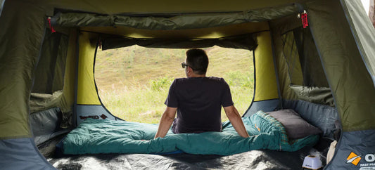 Person relaxing inside an OZtrail Fast Frame tent, sitting on a sleeping bag and looking out toward a grassy hillside.