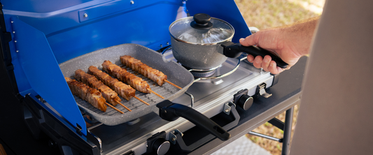 Skewers cooking on a grill pan beside a pot on a portable camping stove, with a hand holding the pot handle.