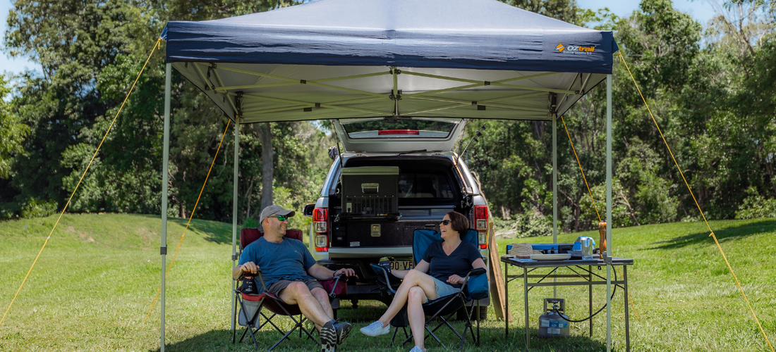 Couple relaxing under a navy Oztrail Deluxe Gazebo 3.0 beside their open 4WD at a sunny grassy campsite.