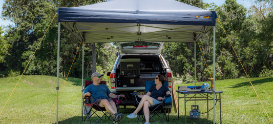 Couple relaxing under a navy Oztrail Deluxe Gazebo 3.0 beside their open 4WD at a sunny grassy campsite.