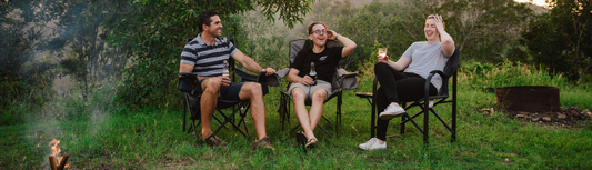 Three friends sitting in camping chairs around a fire pit on grassy hills, laughing and enjoying drinks at sunset.