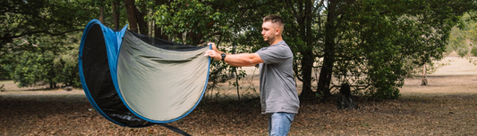 Camper unfolding an Oztrail Pop Up Pod Tent in a bushland campsite.
