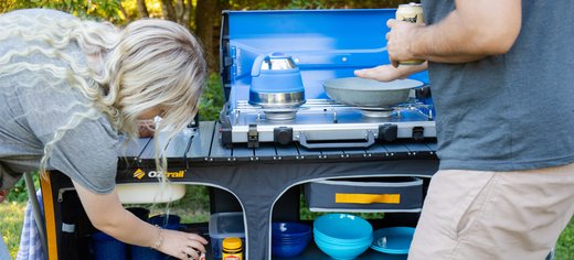 Campers cooking at a portable camp kitchen with a gas stove, cookware, and storage shelves outdoors.