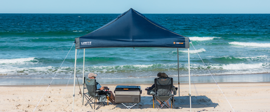 Two people relaxing under a navy Blockout Lumos 3.0 Gazebo on the beach with waves in the background.