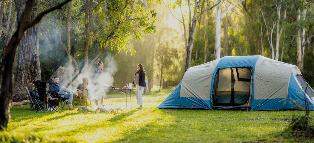 Campers relaxing by a smoky campfire beside a spacious Tasman 10P tent set up in a bright forest clearing.