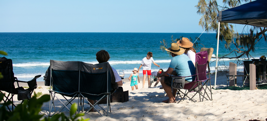 Campers seated in OZtrail chairs on the beach watching children play by the shoreline under a shade gazebo.