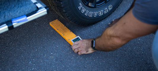 Person placing a Digital Weight Control Scale under a vehicle tyre on asphalt.