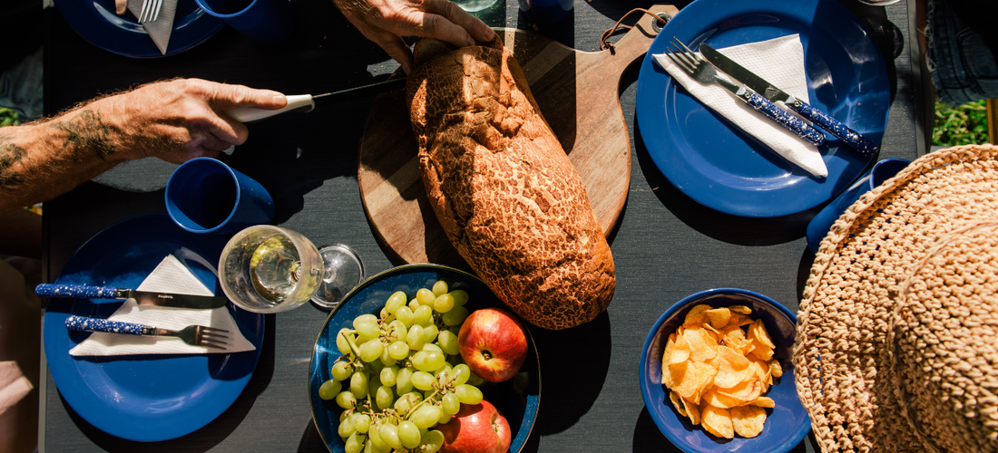 Top-down view of a picnic table with bread, fruit, chips and blue camping plates as someone slices a loaf.