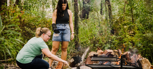 Two women cooking over a campfire in bushland, tending food on a grill surrounded by logs and flames.