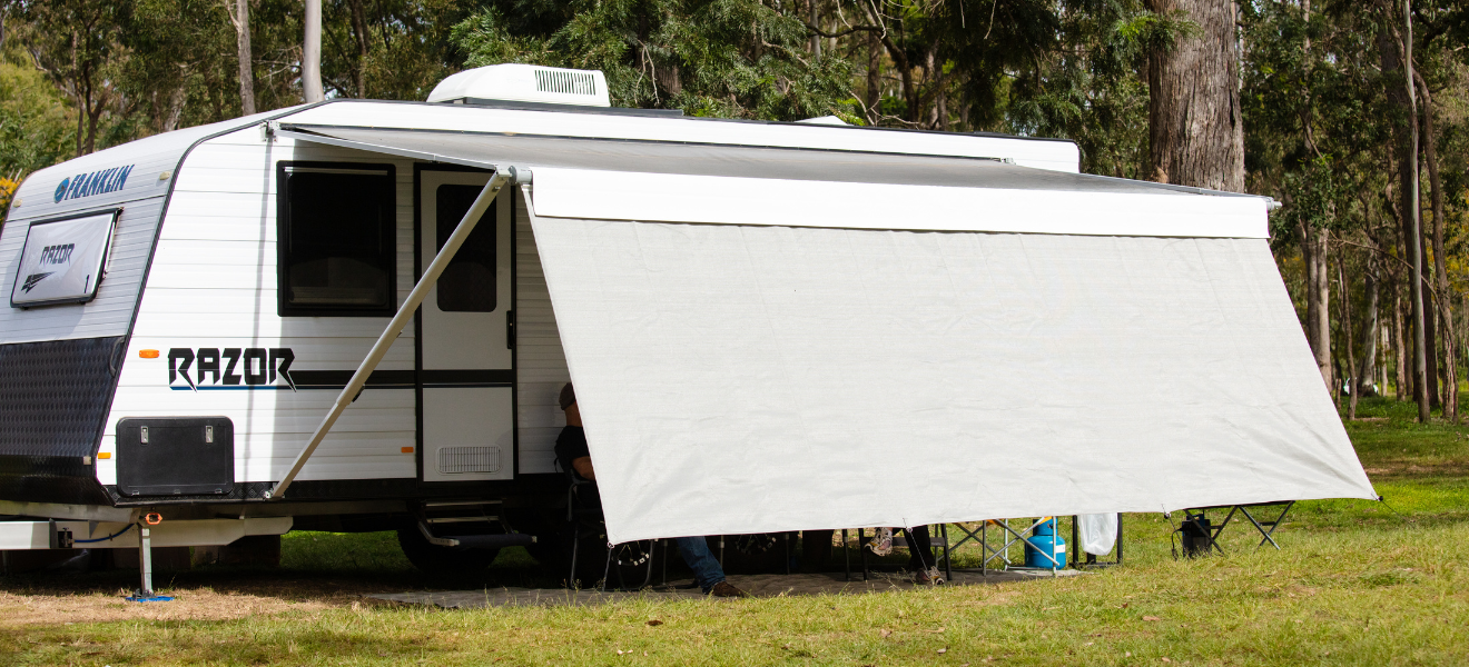 White caravan with awning set up in a grassy campsite, with a person seated underneath in a wooded area.