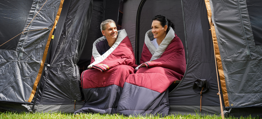 Couple wrapped in Kingsford Double Sleeping Bags sitting by tent in outdoor setting