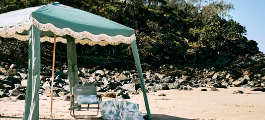 A beach scene features a green cabana, a chair, and beach bags set up on the sand. The background includes a rocky hillside and the ocean.