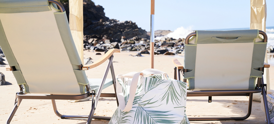 A beach scene features a green cabana, a chair, and beach bags set up on the sand. The background includes a rocky hillside and the ocean.