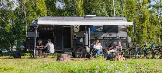 Family relaxing beside an off-road caravan with awning extended, campfire cooking, bikes parked, and forest campsite setting