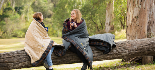 Two women in sleeping bags sit on a log, holding mugs and smiling in a forest setting.