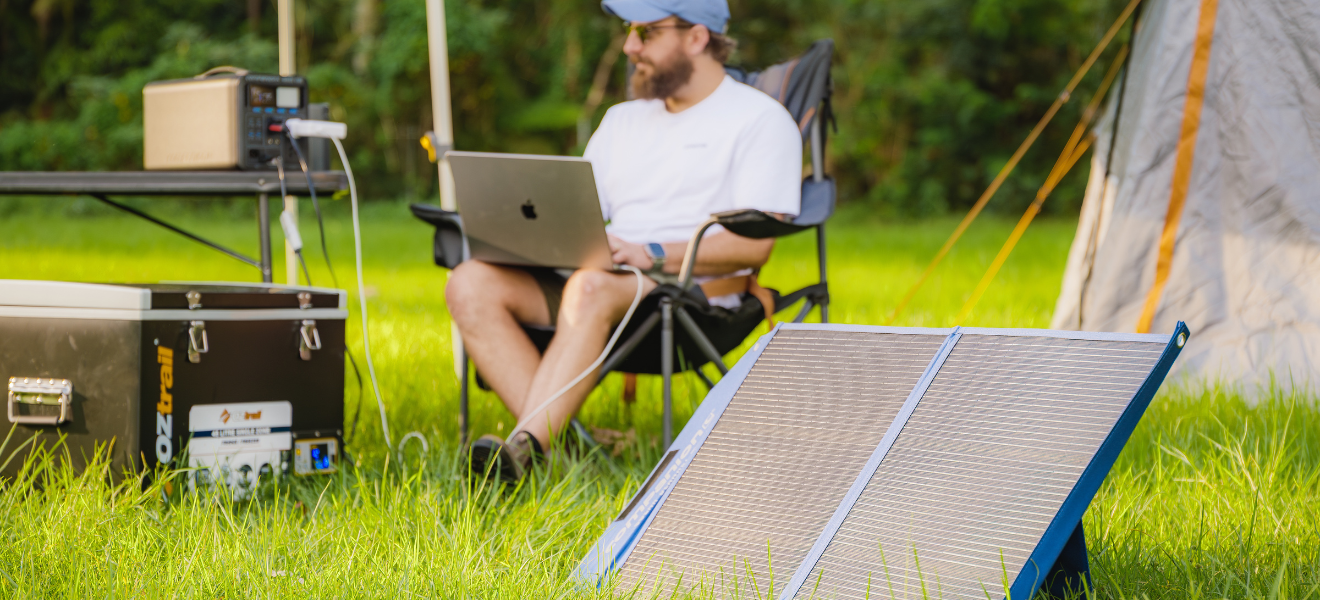 A bearded man in a cap works on a laptop beside the Companion 100W Solar Blanket and camping gear.