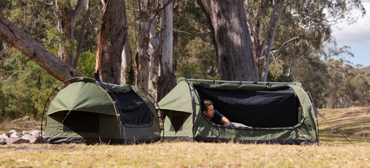 Here's alt text for both, each under 120 characters: Image 1: Two green OZtrail dome swags set up among gum trees, a man resting inside one with the mesh door open.