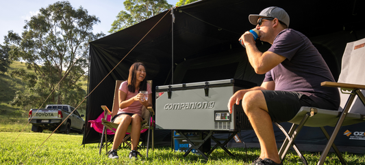 A man and woman relax in camping chairs beside a tent with the Companion 100L Dual Zone Fridge/Freezer, a pickup parked on the hill behind them.