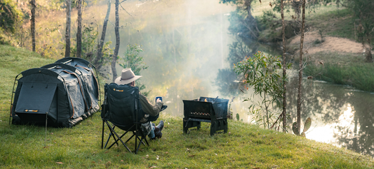 A person sits in a camping chair beside a river, holding a mug and looking towards a smoky campfire.