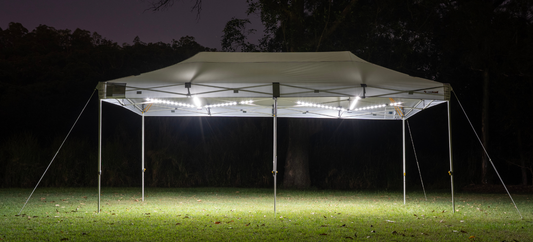 A white Commercial Lumos Gazebo illuminated at night in a grassy, wooded area. Lights beneath the canopy cast a glow.