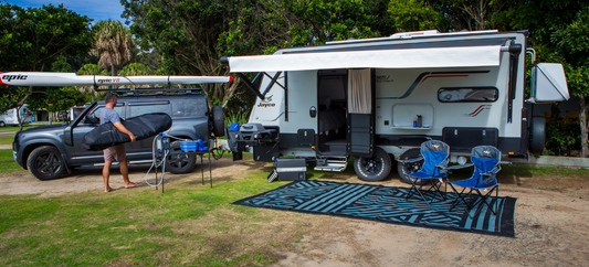 Man carries surfboard bag beside caravan, SUV with kayak, and Caravan Recycled Ground Mat 4M x 2.4M at campsite