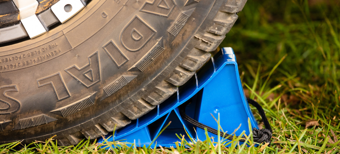 Blue wheel chock secured under a vehicle tyre on grass, helping prevent rolling and improve campsite stability.