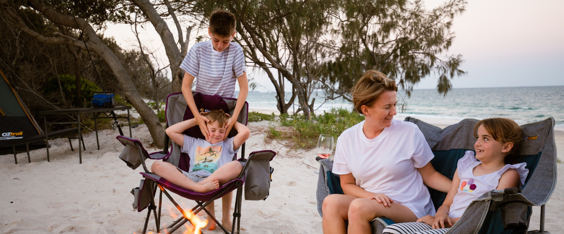 Two kids and their mother sitting in camping chairs on a sandy beach near a small fire, smiling and talking.”