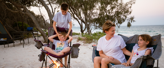 Two kids and their mother sitting in camping chairs on a sandy beach near a small fire, smiling and talking.”