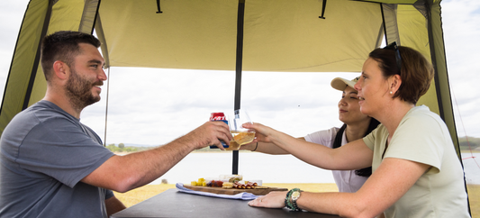 ChatGPT said:  Three campers clinking drinks at a table under a gazebo, with a lakeside view and shared snacks.