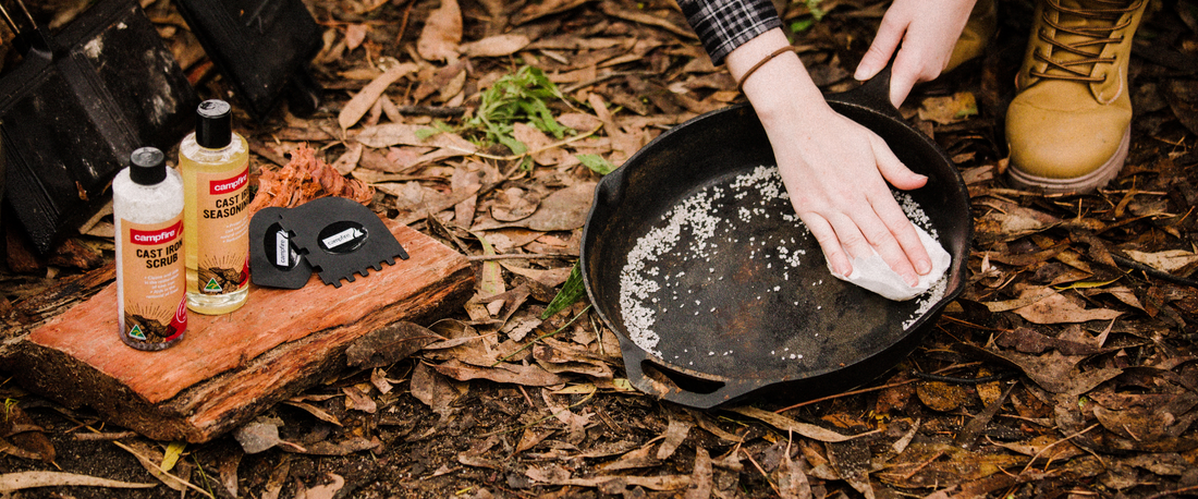 Person scrubbing a cast iron pan with salt next to Campfire cast iron cleaning products on the ground among leaves.