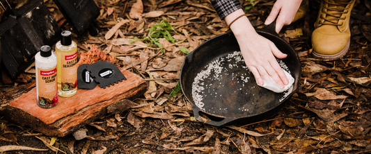 Person scrubbing a cast iron pan with salt next to Campfire cast iron cleaning products on the ground among leaves.